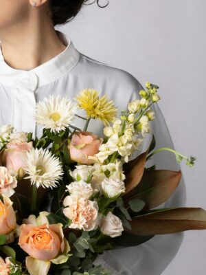 Bouquet Luce, mazzo di fiori incartato con rose pesca, garofani chiari, violaciocca crema, germini spider bianchi e gialli, roselline ramificate chiare e verde di stagione. Ragazza con camicia bianca tiene in mano un bouquet di fiori con incarto Frida's