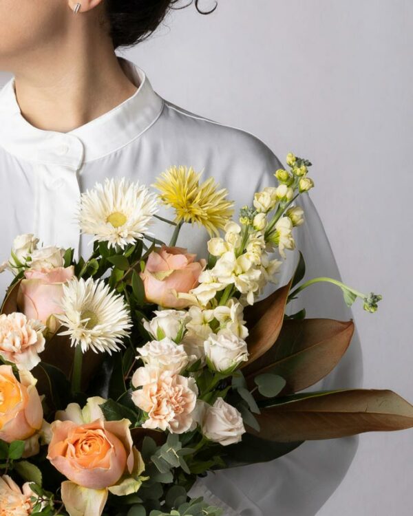 Bouquet Luce, mazzo di fiori incartato con rose pesca, garofani chiari, violaciocca crema, germini spider bianchi e gialli, roselline ramificate chiare e verde di stagione. Ragazza con camicia bianca tiene in mano un bouquet di fiori con incarto Frida's